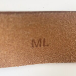 May include: Close-up of a brown leather tag with the letters "ML" embossed in black.
