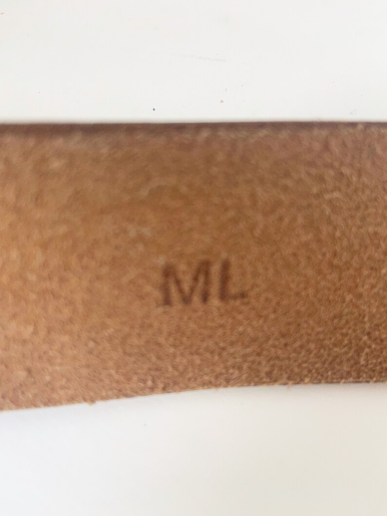 May include: Close-up of a brown leather tag with the letters "ML" embossed in black.