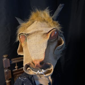 Könnte beinhalten: Eine Maske aus Jute mit bemalten Details, die ein kuhähnliches Gesicht mit großen Augen, einer Schnauze und Zähnen darstellt. Die Maske hat Kunstfell um die Stirn und schwarze Hörner. Konzipiert für Kostüm- oder Theaterzwecke.