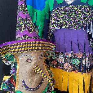 May include: A whimsical Halloween costume featuring a witch hat and a mask with a long nose. The hat is purple and black with a yellow brim. The mask is burlap with a smiling face. The costume includes a top with colorful fabric and fringe.