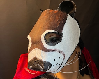 May include: A brown and white animal mask with a long snout and whiskers. The mask is made of paper mache and has a painted finish.