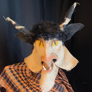 May include: A handmade mask with a burlap face, yellow eyes, and a pink nose. The mask has black fur and horns with black and tan stripes. The mask is worn with an orange and black checkered scarf.
