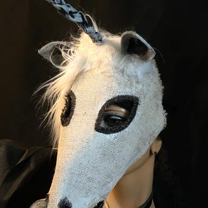 May include: A white burlap unicorn mask with black accents around the eyes and muzzle. The mask has a white mane and a twisted horn with black and white patterns. The mask is on a mannequin head against a black background.