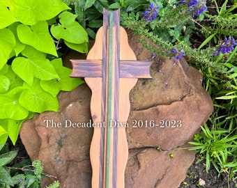 May include: A wooden cross with a green and brown striped design. The cross is sitting on a large brown rock with green plants and purple flowers in the background. The text "The Decadent Diva 2016-2023" is visible on the rock.