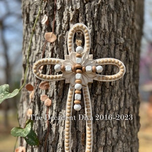 May include: A white wooden cross with a brown beaded border. The cross has a carved floral design in the center.
