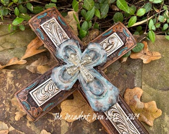 May include: A rustic wooden cross with a turquoise and brown distressed finish. The cross features a silver-toned metal overlay with intricate designs and a smaller cross with rhinestones in the center. The cross is surrounded by brown leaves.