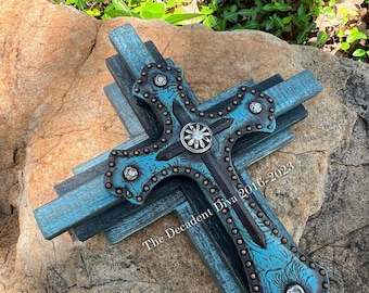 May include: A turquoise blue wooden cross with a black and copper center. The cross is decorated with rhinestones and has a distressed finish. The text "The Decadent Diva 2016-2023" is visible in the lower right corner.