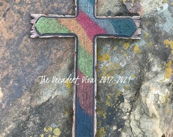 May include: A colorful beaded cross with a wooden frame. The beads are arranged in a diagonal pattern, creating a rainbow effect. The cross is set against a rocky background. The text "The Decadent Diva 2017-2021" is visible at the bottom of the cross.