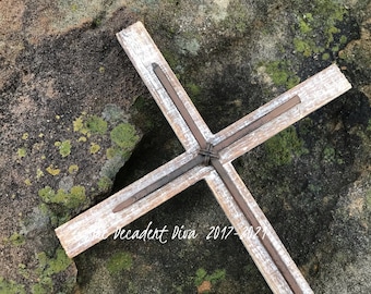Peut inclure: Une croix composée de deux lattes de bois blanchies et usées, assemblées au centre par un fil métallique sombre, avec quatre longs clous en fer rouillé disposés le long de chaque bras en bois. Le texte "Decadent Diva 2017-2021" est visible.