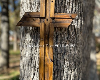 Può includere: Una croce di legno con una finitura rustica, montata su un tronco d'albero. La croce è realizzata con più pezzi di legno, creando un effetto a strati. Il testo "The Decadent Diva 2016-2023" è visibile nella parte inferiore della croce.