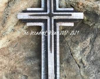 May include: A rustic wooden cross with a distressed white and black paint finish. The cross is decorated with a piece of barbed wire at the center. The text "The Decadent Diva 2017-2021" is visible at the bottom of the cross.