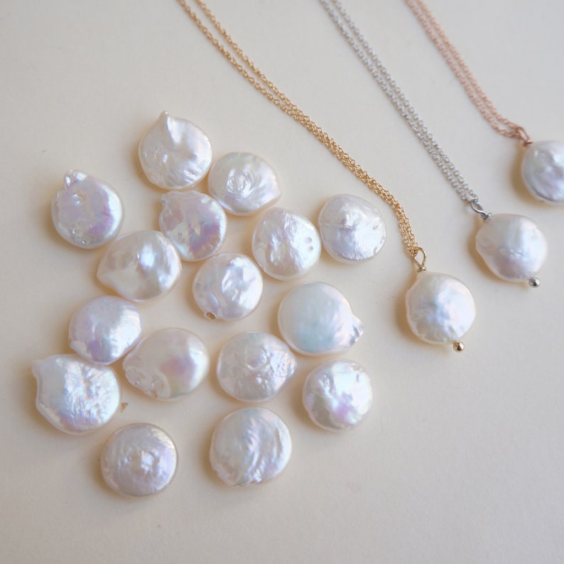 May include: A collection of white, round, pearl-like beads, some of which are strung on gold and silver chains. The beads are scattered on a light beige surface.