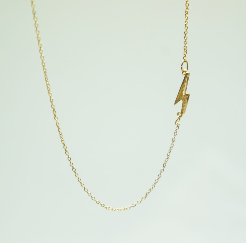 May include: A delicate gold-colored necklace featuring a lightning bolt pendant. The pendant is a stylized representation of a lightning bolt. The chain is thin and appears to be made of small, interconnected links. The necklace is displayed against a plain white background.