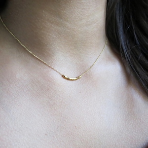 May include: A delicate gold necklace featuring a curved bar pendant with small, cylindrical beads. The necklace is displayed on a person's neck, showcasing its minimalist design and subtle elegance. The chain is fine and the pendant is approximately 1 inch in length.