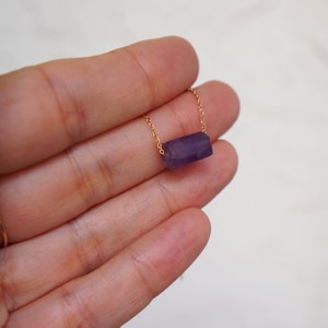 May include: A delicate gold chain necklace with a purple amethyst stone pendant. The stone is a rectangular shape and has a natural, raw look.