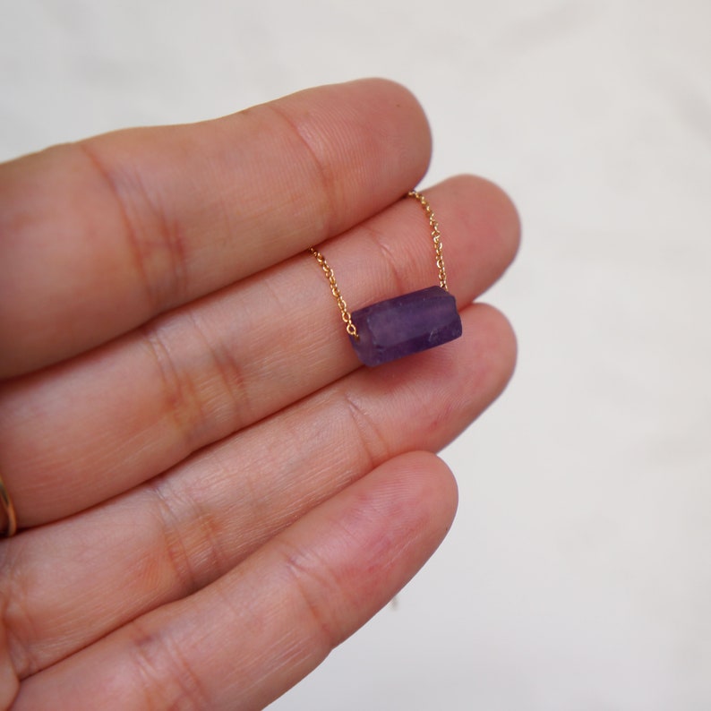 May include: A delicate gold chain necklace with a purple amethyst stone pendant. The stone is a rectangular shape and has a natural, raw look.