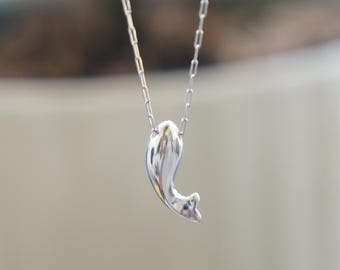 May include: A silver-colored necklace with a pendant shaped like a stylized dolphin. The pendant is smooth and reflective, hanging from a delicate chain. The background is blurred, emphasizing the jewelry.