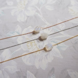 May include: Three delicate necklaces with a single, raw moonstone pendant each. The necklaces are shown on a white floral background. The necklaces are available in gold, silver, and rose gold.