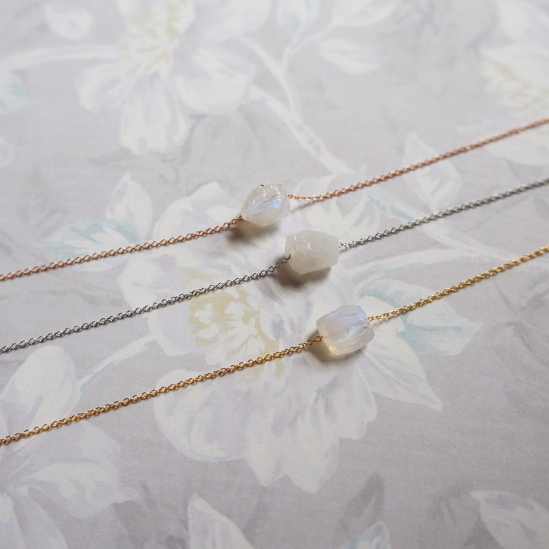 May include: Three delicate necklaces with a single, raw moonstone pendant each. The necklaces are shown on a white floral background. The necklaces are available in gold, silver, and rose gold.