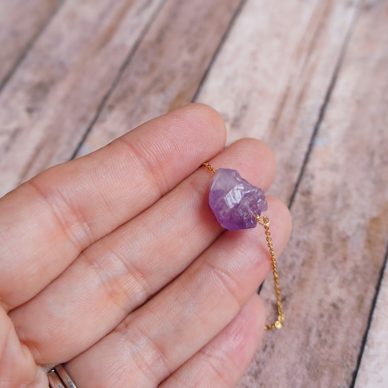 Puede incluir: Un delicado collar de cadena dorada con un colgante de cristal de amatista en bruto. El cristal es de color morado intenso y tiene una forma natural irregular.