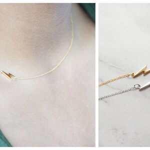 May include: A delicate gold necklace featuring a lightning bolt pendant. The necklace is shown on a person's neck, and a second image shows the necklace in gold and silver. The chain is thin and the lightning bolt is small.