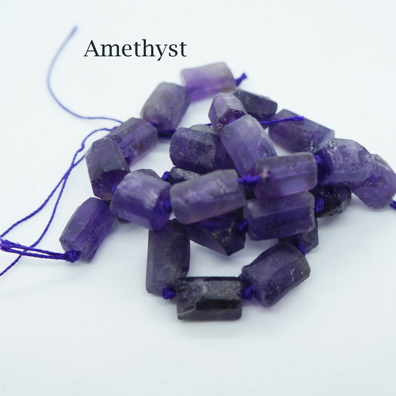 May include: A strand of purple amethyst gemstone beads, each bead is a rectangular shape with rough edges, strung on a purple thread.