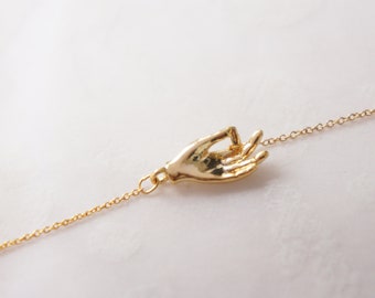 May include: A gold chain necklace with a gold hand charm in the shape of a hand gesture. The hand is in a relaxed, open position with the fingers slightly curled.