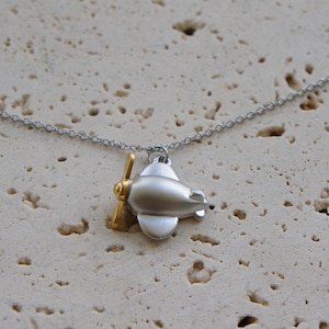 May include: A silver airplane pendant necklace with a gold propeller. The airplane charm is suspended from a delicate silver chain. The necklace is displayed on a textured, light-colored surface.