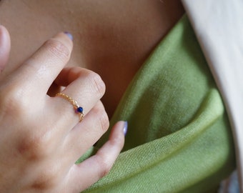 May include: A delicate gold chain ring with a small blue gemstone. The ring is worn on the finger of a person wearing a green shirt.
