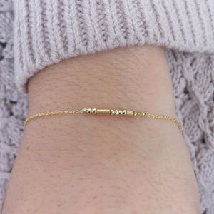 May include: A delicate gold bracelet with a series of small, cylindrical beads and chain links. The bracelet is worn on a wrist, with a light-colored sweater in the background. The bracelet is a piece of jewelry.