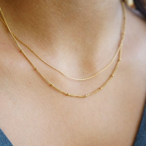 May include: Two gold necklaces layered on a person's neck. The top necklace is a delicate chain with small beads. The bottom necklace is a thicker chain with larger beads.