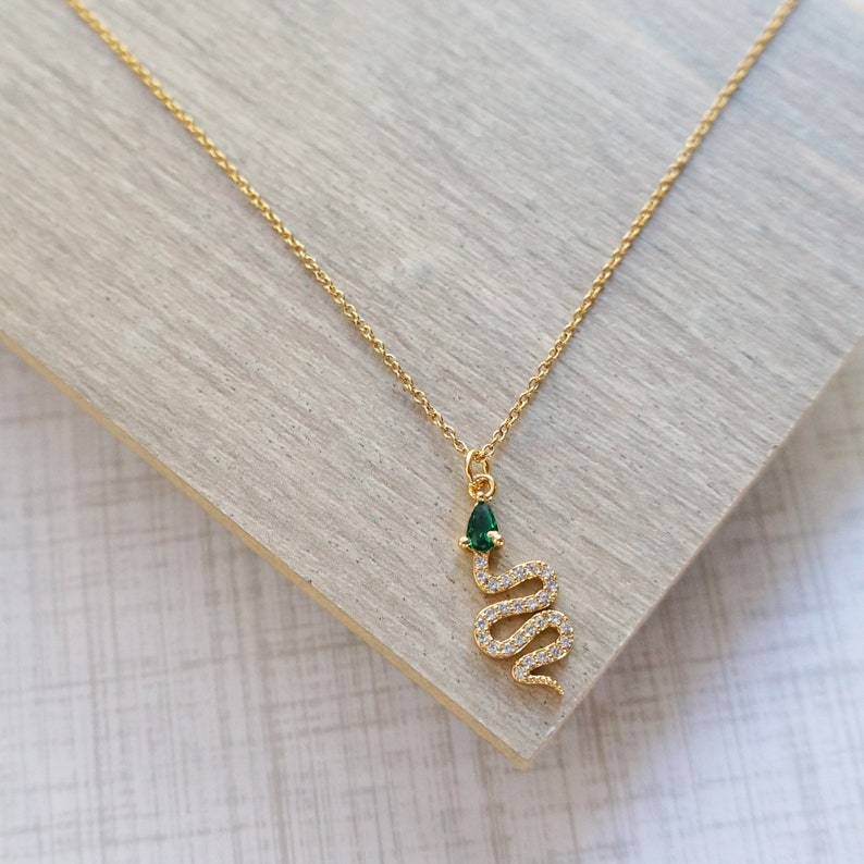 May include: A gold chain necklace with a snake pendant. The snake is encrusted with clear rhinestones and has a green gemstone for a head.