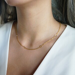 May include: A delicate gold-colored necklace featuring small heart-shaped charms spaced along a fine chain. The necklace is worn against a white top, highlighting the jewelry's design. The chain is approximately 18 inches long.