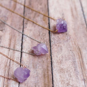 Puede incluir: Tres delicados collares con colgantes de cristal de amatista sobre un fondo de madera. Cada collar presenta un estilo de cadena diferente, incluyendo plata, oro y oro rosa.