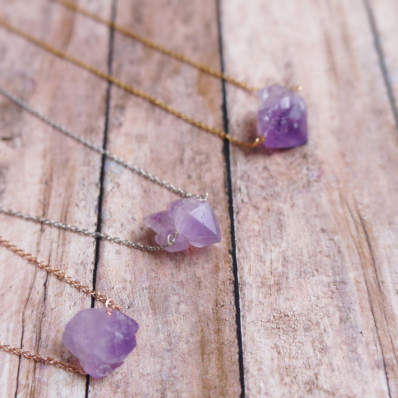 Puede incluir: Tres delicados collares con colgantes de cristal de amatista sobre un fondo de madera. Cada collar presenta un estilo de cadena diferente, incluyendo plata, oro y oro rosa.