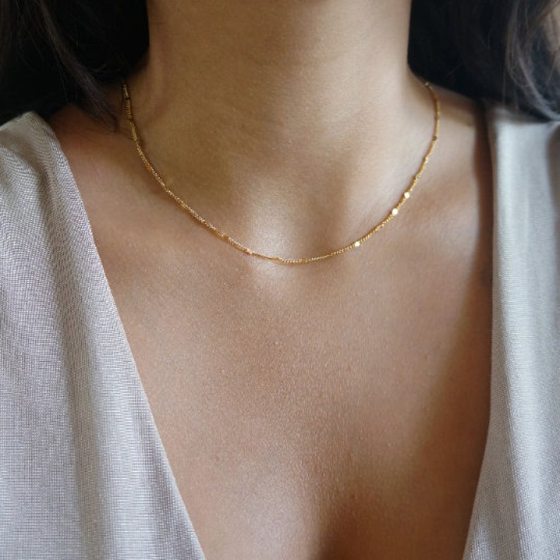 May include: A delicate gold necklace with a fine chain and small, evenly spaced circular accents. The necklace is worn around the neck, resting against the skin. The jewelry is a simple, elegant accessory.