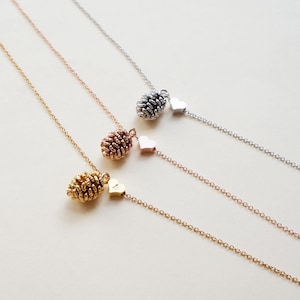 May include: Three delicate necklaces with gold, rose gold, and silver chains. Each necklace features a small pine cone charm and a heart charm.