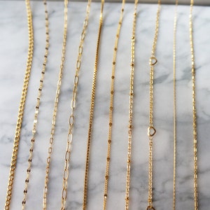 May include: A collection of delicate gold necklaces displayed on a marble surface. The necklaces feature various chain styles, including curb, oval link, box, and beaded designs. Some necklaces have heart-shaped pendants.
