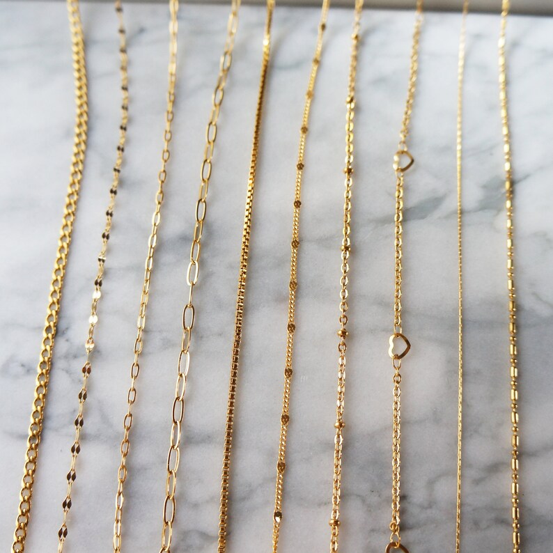 May include: A collection of delicate gold necklaces displayed on a marble surface. The necklaces feature various chain styles, including curb, oval link, box, and beaded designs. Some necklaces have heart-shaped pendants.