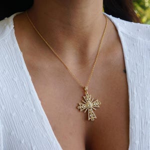 以下が含まれることがあります： 金色のペンダントネックレス。十字架のデザインで、小さな透明な宝石がちりばめられ、詳細で装飾的な模様があります。白い背景にネックレスが表示されています。