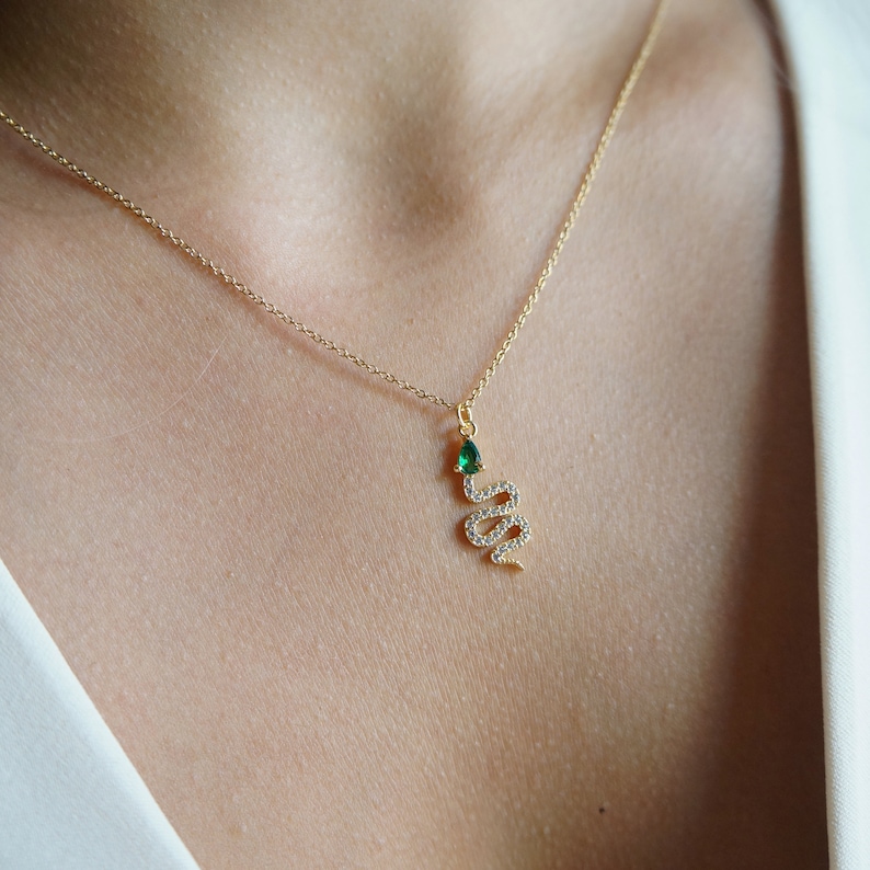 May include: A gold chain necklace with a snake pendant. The snake is adorned with small clear stones and has a green gemstone for a head.