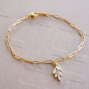 May include: A gold chain bracelet with a delicate pendant featuring a cluster of clear, marquise-shaped stones. The pendant resembles a stylized leaf.