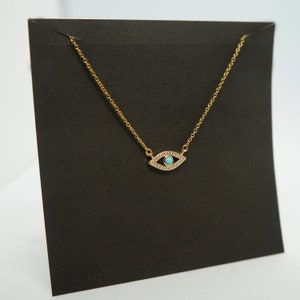 May include: A gold chain necklace with a small turquoise blue eye charm. The eye is surrounded by clear rhinestones.