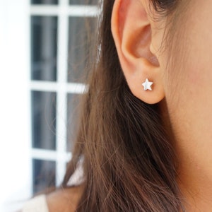 K&ouml;nnte beinhalten: Nahaufnahme eines Ohrs mit einem kleinen, silbernen sternf&ouml;rmigen Ohrstecker. Der Ohrring ist ein schlichtes, minimalistisches Design. Die Person hat lange, braune Haare.