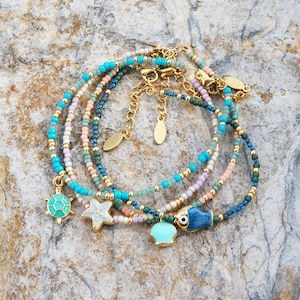 May include: Multi-strand beaded bracelet with turquoise, blue, and pink beads. The bracelet features gold charms of a turtle, starfish, and a fish with an evil eye.