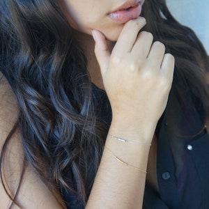 May include: Two delicate bracelets, one silver and one gold, adorn a wrist. The silver bracelet features small beads, while the gold bracelet has a bar design. The bracelets are set against a dark background.