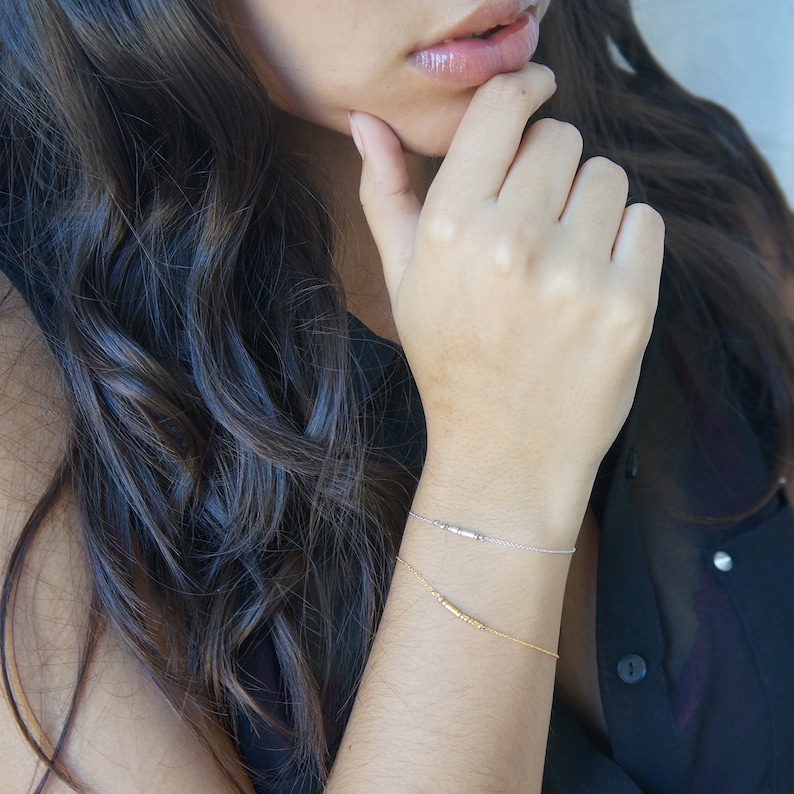 May include: Two delicate bracelets, one silver and one gold, adorn a wrist. The silver bracelet features small beads, while the gold bracelet has a bar design. The bracelets are set against a dark background.