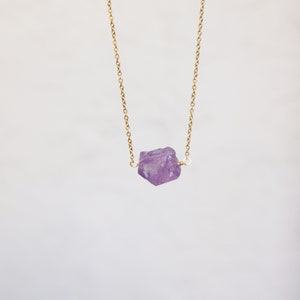 Puede incluir: Un delicado collar de cadena dorada con un colgante de cristal de amatista en bruto. El cristal es de color p&uacute;rpura claro y tiene una forma natural irregular.