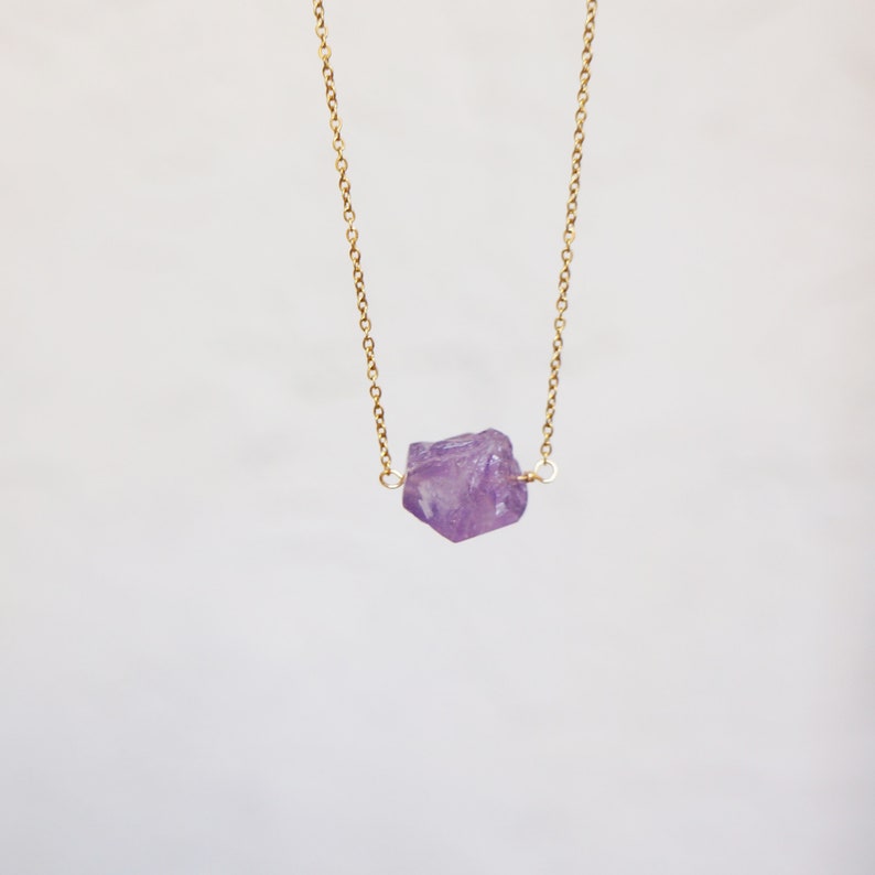 Puede incluir: Un delicado collar de cadena dorada con un colgante de cristal de amatista en bruto. El cristal es de color p&uacute;rpura claro y tiene una forma natural irregular.