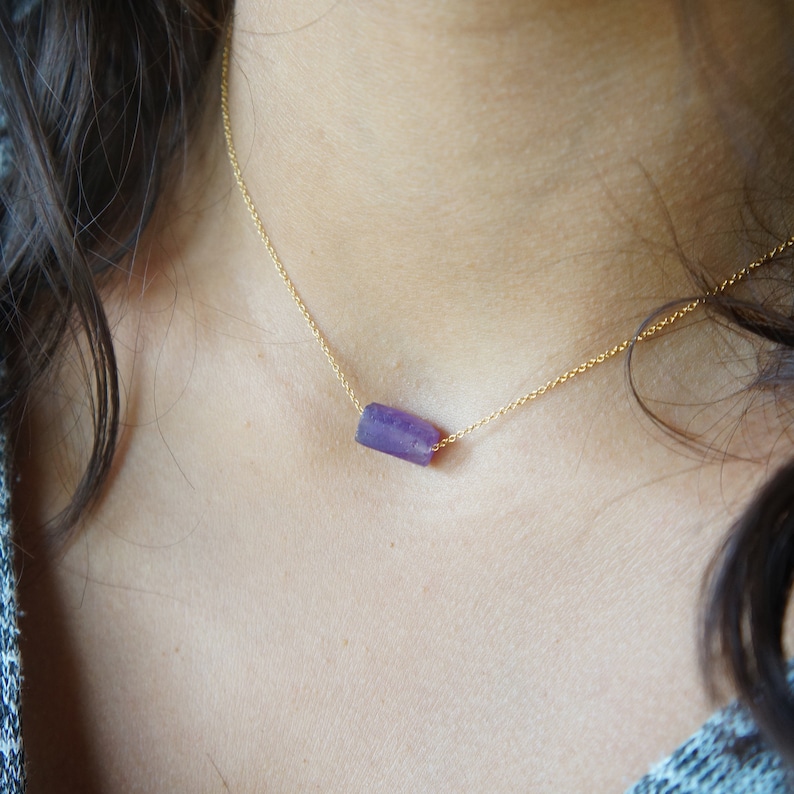 May include: A gold chain necklace with a purple gemstone pendant. The pendant is a rectangular shape with a rough, natural texture.